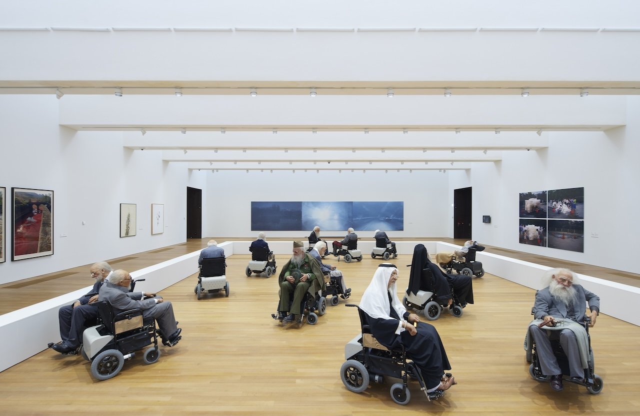Installation view of thirteen sculptures of elderly people in electric wheelchairs. Each elderly person is dressed in a different attire while the wheelchairs move in an area enclosed by white fences. The installation is surrounded by artworks mounted on the white walls of the gallery.